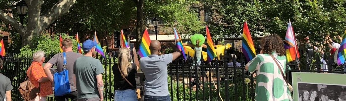 A copy of the original hero image for the Stonewall National Monument website showing people standing by a fence waving pride flags as a performer in a gorgeous green wig dances.